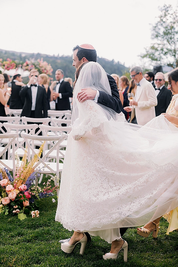 Ceremony moment as bride and groom hug, groom in tuxedo with kippah beside white chairs on an outdoor lawn with hills behind