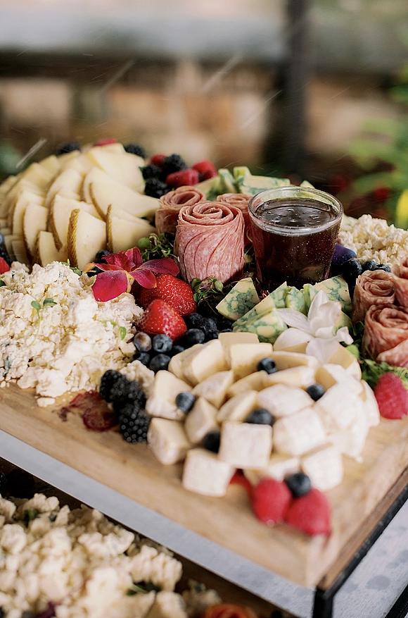 Charcuterie board at wedding cocktail hour with sliced cheeses, salami rose, berries, pear, jam, and edible flowers on a wood board outdoors