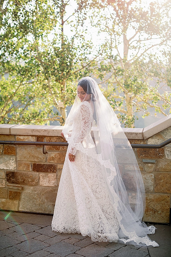 Bridal portrait of a bride in a long sleeve lace wedding dress with cathedral veil and earrings, standing by a sunlit stone wall patio