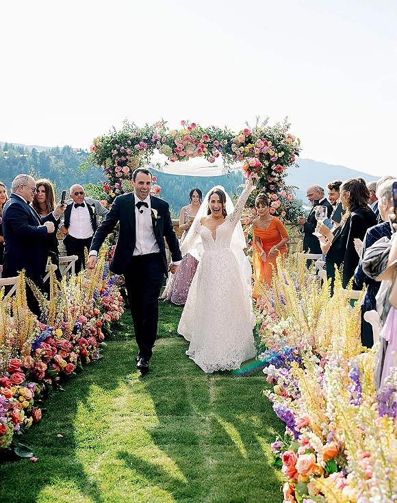 Wedding recessional as bride and groom walk down aisle hand in hand under floral arch, guests cheering with mountain view behind