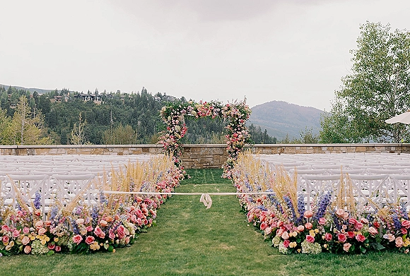 Ceremony aisle decor with outdoor ceremony aisle flowers lining a lawn, leading to a rose floral arch with ribbon, mountains beyond