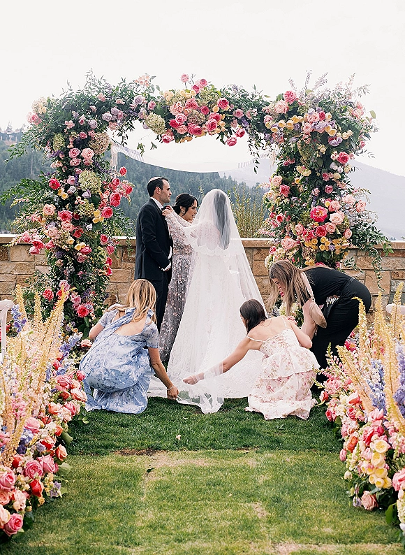 Ceremony moment at an outdoor wedding ceremony with bride and groom under a floral arch, mountain landscape behind and veil flowing
