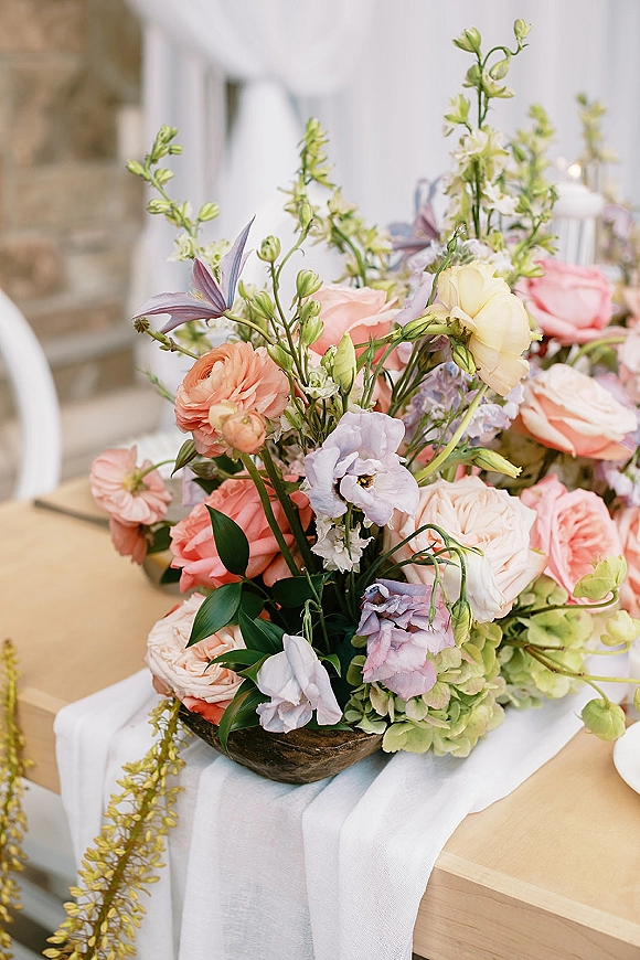 Wedding centerpiece in a wooden bowl with garden roses, ranunculus, greenery, and taper candles on a linen runner table with stone wall backdrop