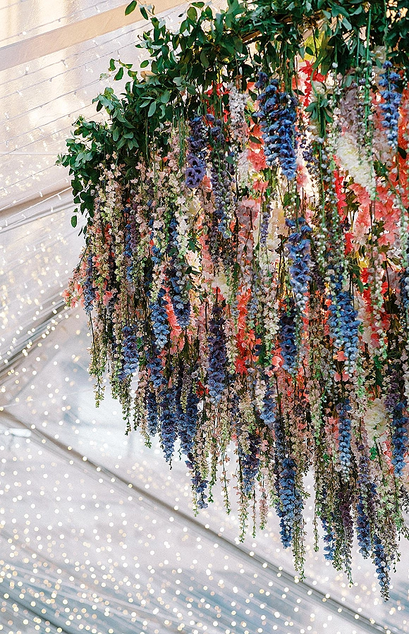 Hanging floral installation with cascading greenery and a floral chandelier, accented by string lights under a white tent ceiling