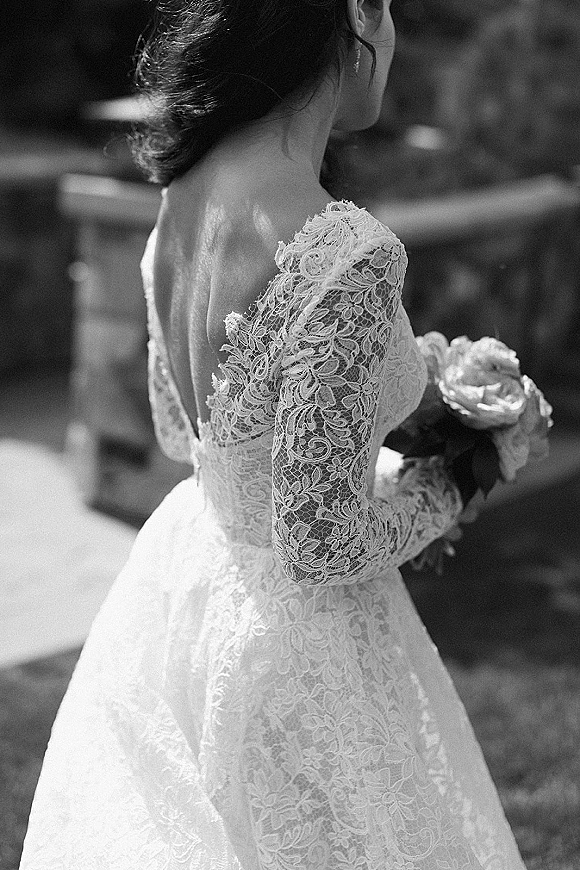 Wedding dress close-up of a lace wedding dress with long sleeves and open back, bouquet held behind her on a sunlit garden path