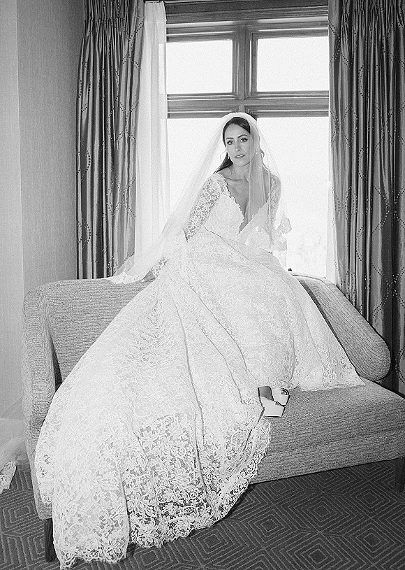 Bridal portrait in window light of a bride in a long-sleeve lace gown and cathedral veil, seated on a chaise in a hotel room