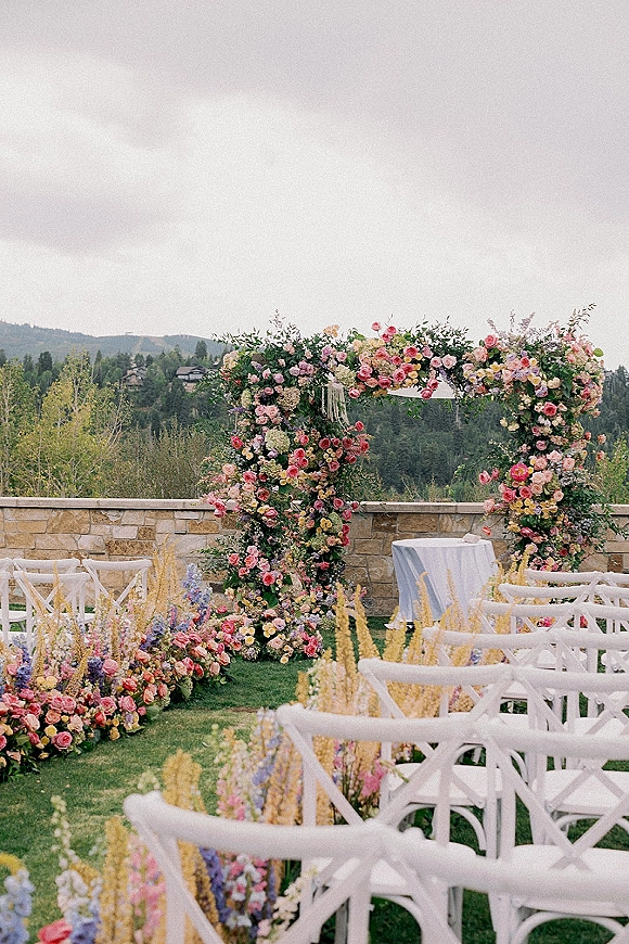 Ceremony setup with an outdoor wedding ceremony setup, featuring a floral arch of roses and greenery beside white chairs and mountains