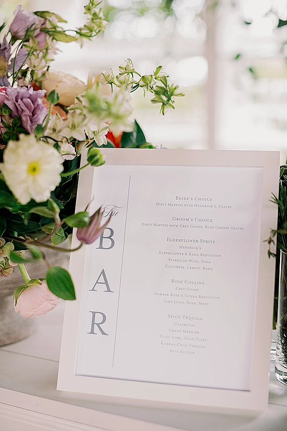 Wedding bar menu card with classic typography beside a floral arrangement and greenery in a vase on a white tabletop in window light