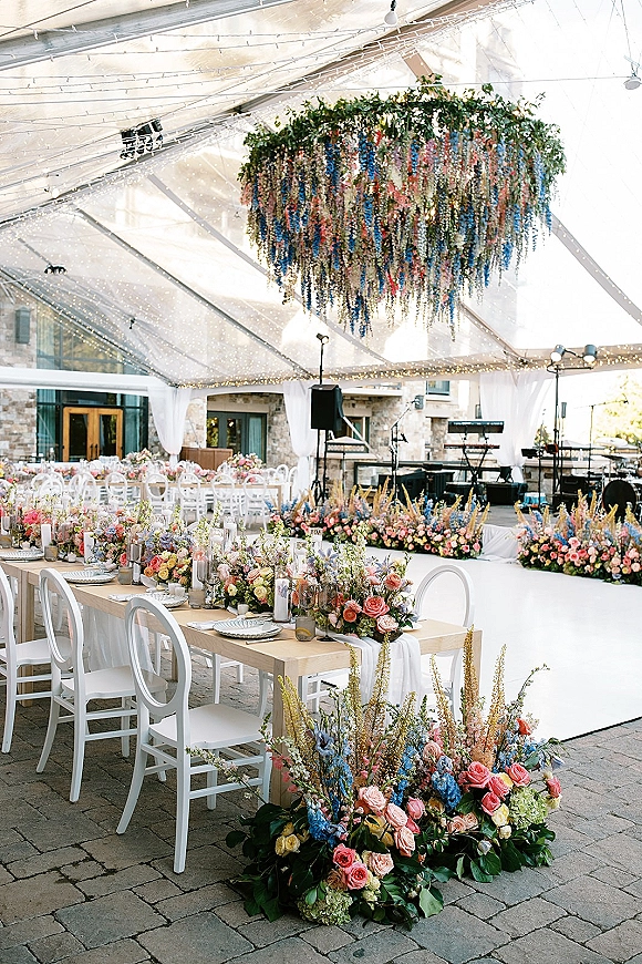 Reception tablescape with wedding head table decor on long farm tables, pastel florals, candles and glassware under a clear tent with string lights