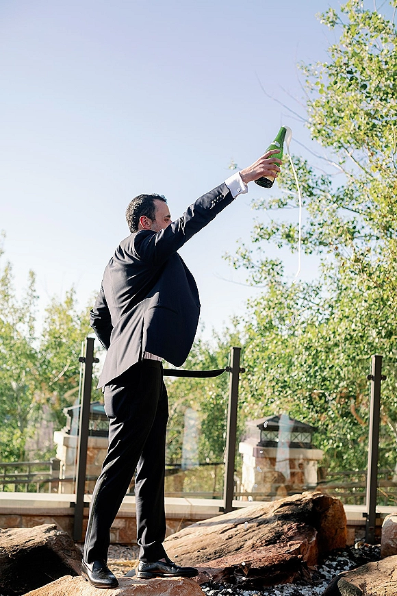 Groom champagne pop in a black tux, champagne spray photo bursting from the bottle on a terrace with glass railing and blue sky