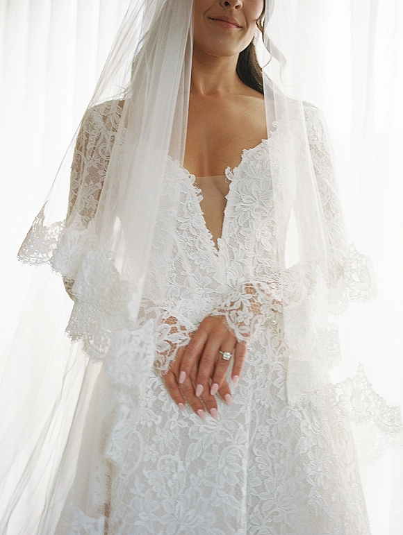 Bridal gown with lace wedding dress details, long sleeves and deep V neckline, shown in bright window light beside sheer curtains