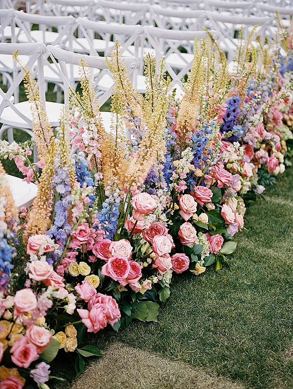 Ceremony aisle florals with wedding aisle flowers of roses, delphinium, snapdragons and greenery lining white folding chairs on grass lawn