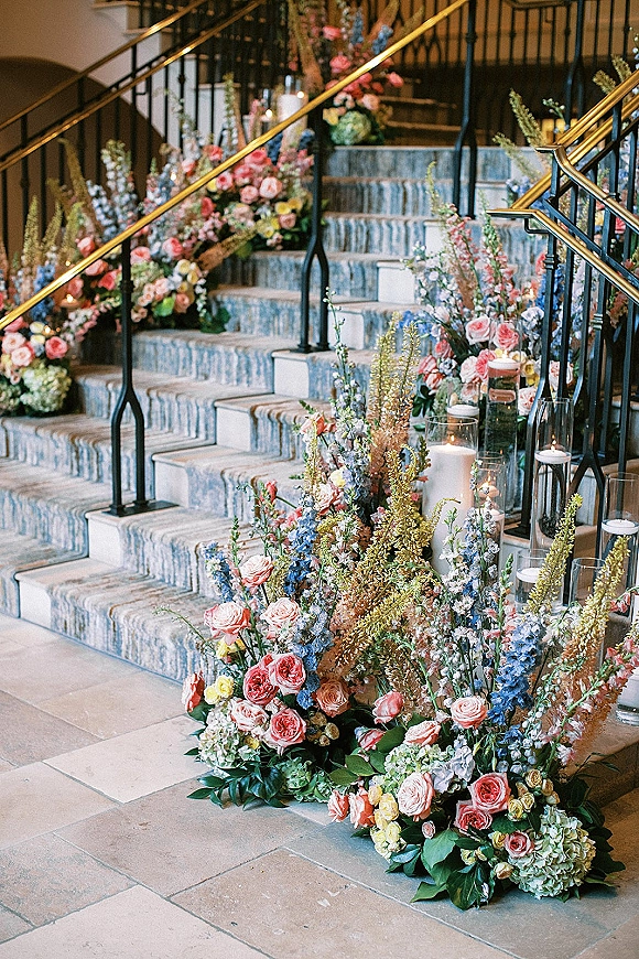 Wedding staircase florals with rose and hydrangea ground arrangements and cylinder candles lining a foyer staircase with runner and brass rails
