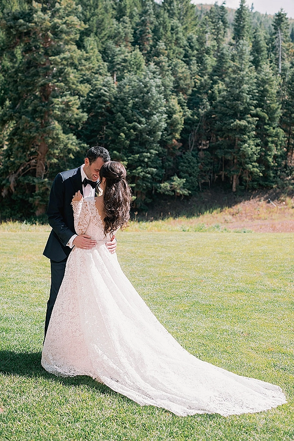 Couple portrait of bride and groom embracing with a forehead touch, her lace long-sleeve gown and train on grass, evergreens behind