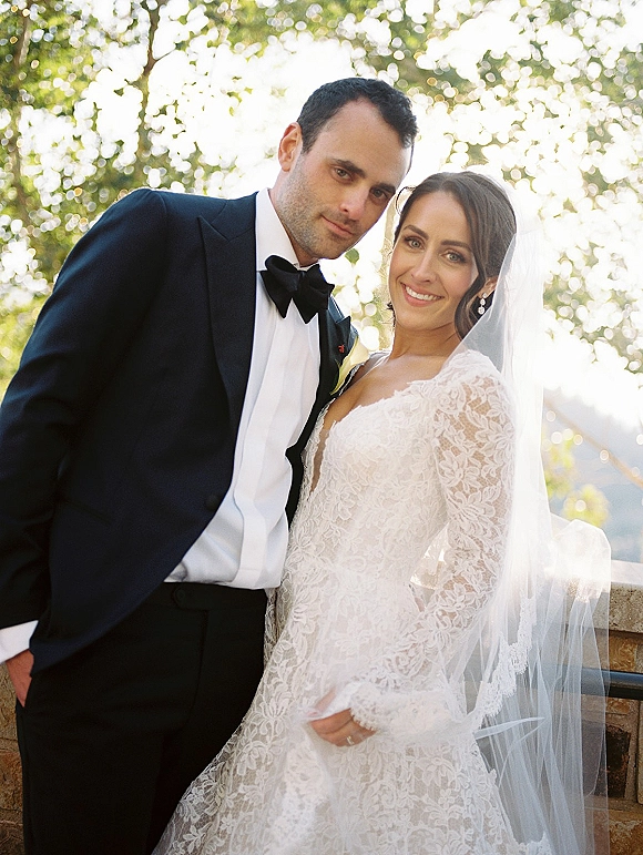 Wedding couple portrait of bride in lace dress and veil with groom in tuxedo and bow tie, smiling by a stone railing in sunlit trees