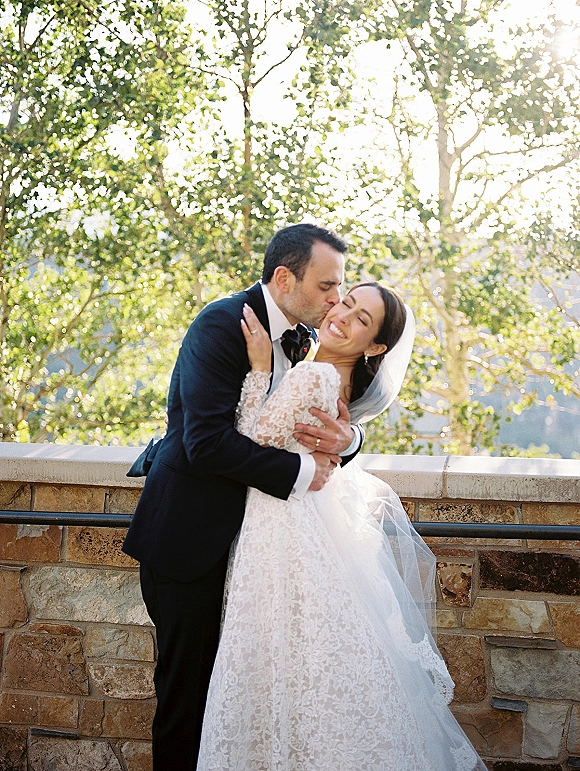 Wedding kiss portrait of groom kissing bride’s cheek as they embrace, lace dress and veil glowing in sun flare by trees and stone wall