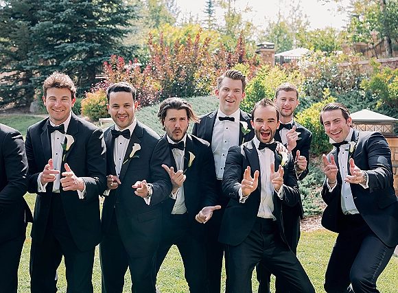 Groomsmen portrait of men in black tuxedos with bow ties and white calla lily boutonnieres, posing on a garden lawn by a stone wall