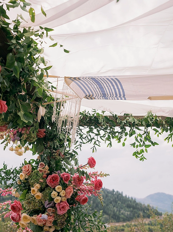 Wedding arch decor with a floral wedding arch of roses, hydrangea, and greenery garland, fabric draping, and fringe with mountains behind