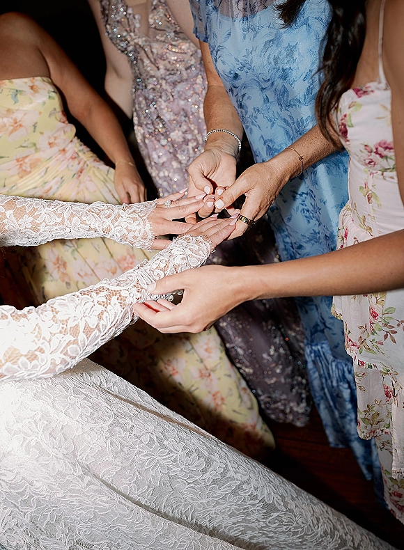Wedding ring exchange close up wedding rings as bridesmaids help slide a band onto the bride’s manicured hand beside lace sleeves in a dark room