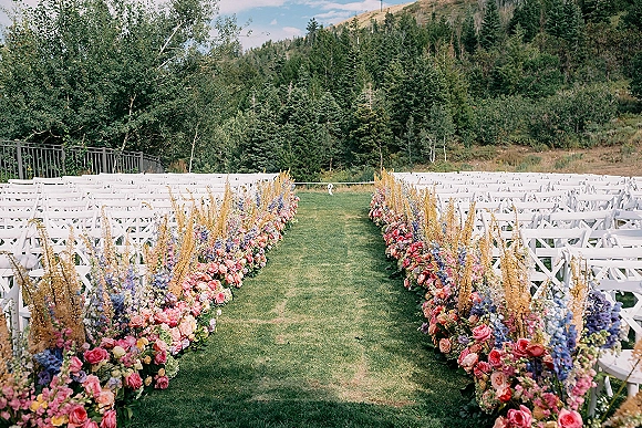 Ceremony aisle decor with a floral lined wedding aisle of pink roses and blue blooms beside white crossback chairs, set against mountains and pines