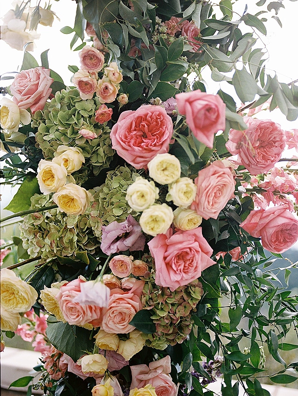 Wedding floral arch with pink and cream roses and green hydrangea accents, lush greenery framing an outdoor landscape under bright sky