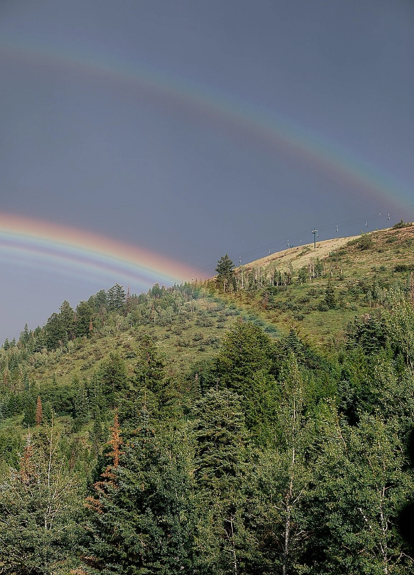 Rainbow landscape with a double rainbow sky arching over an evergreen forest hillside, cloudy storm light, and ski lift cables on the slope