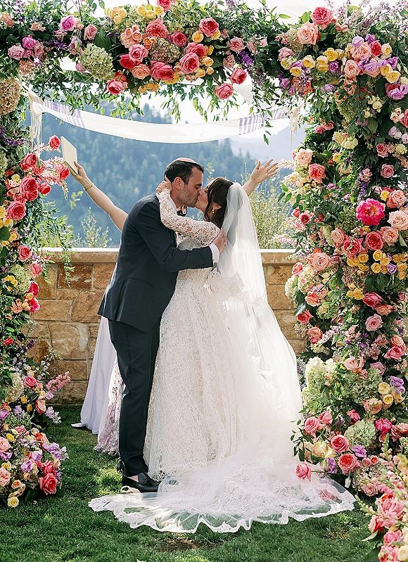 Wedding kiss under a rose floral arch with sheer drapery, bride in lace dress and veil, groom in suit and kippah, mountains behind