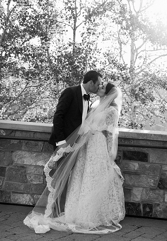Wedding kiss portrait of bride and groom kiss, her long lace veil trailing as sunlight filters through trees by a stone wall walkway
