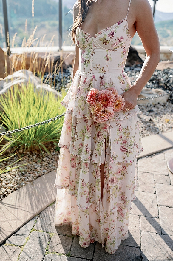 Floral wedding guest dress with a tiered spaghetti-strap skirt, holding a pink bouquet on a sunny stone walkway with mountains behind
