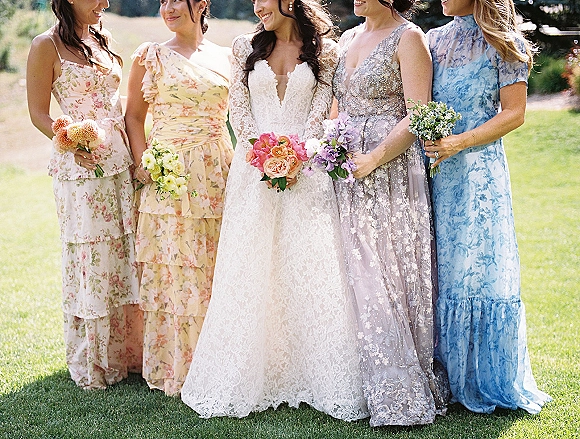 Bride with bridesmaids posing for a bridesmaid group photo, holding peach and purple bouquets on a sunlit lawn with greenery in back