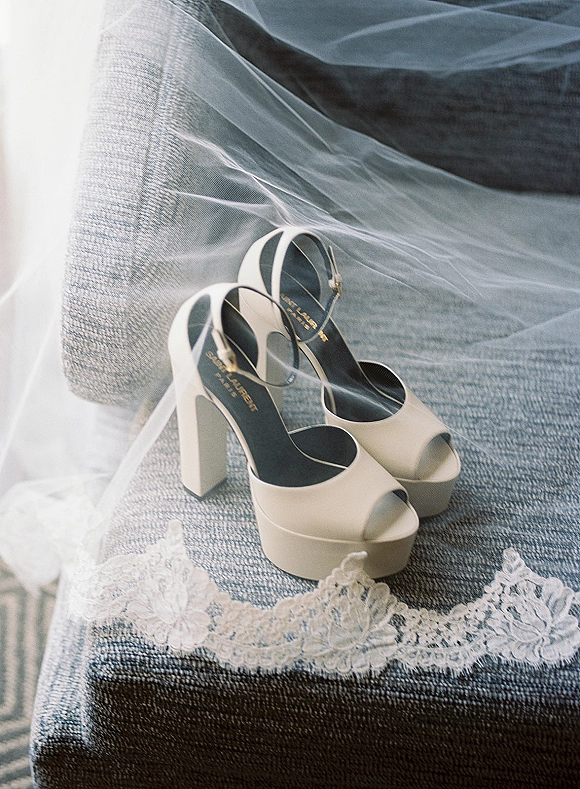 Bridal shoes with white platform wedding heels and ankle straps beside a lace-trim wedding veil on an upholstered chair in a neutral room