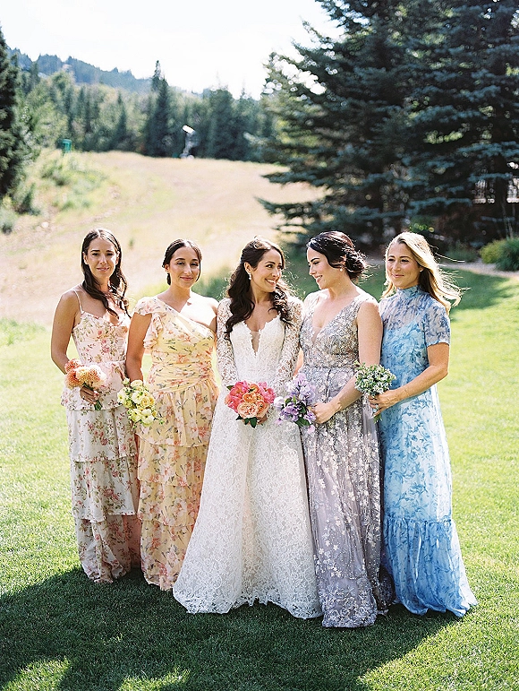 Bridesmaid group portrait with bride centered, holding bouquets in mismatched dresses on a grassy lawn with pine trees and mountains backdrop