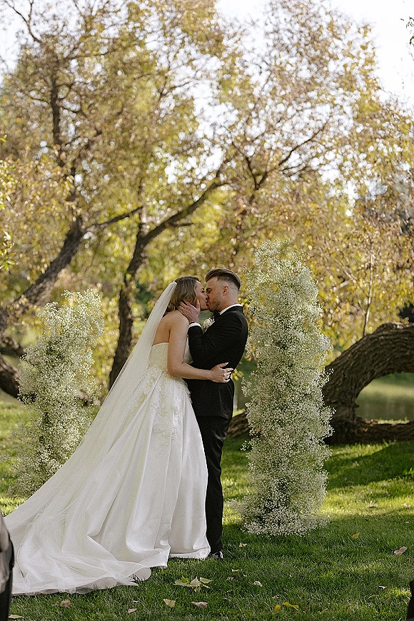 Wedding kiss portrait of bride and groom kissing under a veil, framed by baby's breath floral pillars on a green park lawn