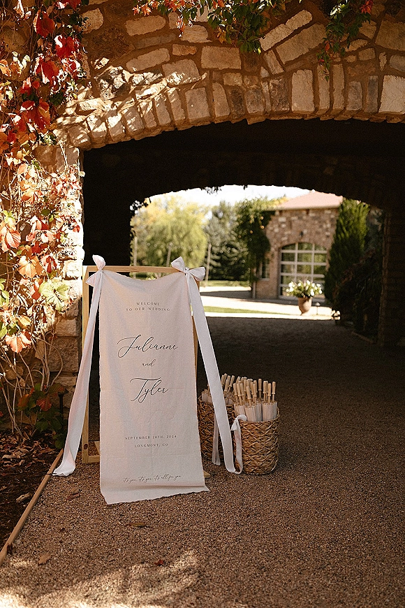 Wedding welcome sign on a wooden stand with ribbon bow and programs in a woven basket beside a sunlit stone archway entrance