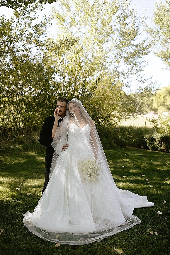 Couple portrait of bride and groom embracing on a sunlit lawn, her cathedral veil draped and white bouquet held beside his tuxedo
