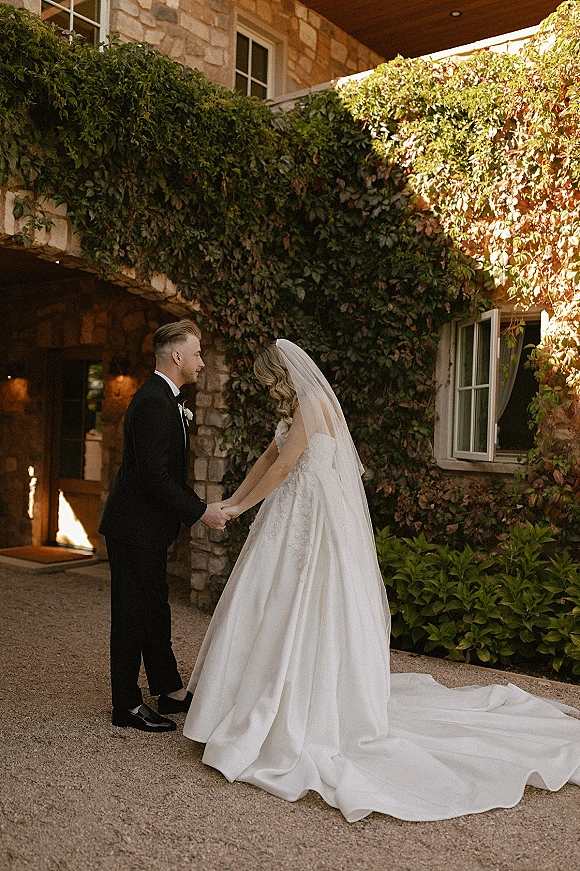 First look moment as bride in wedding dress and cathedral veil holds hands with groom in tux by an ivy-covered stone courtyard wall
