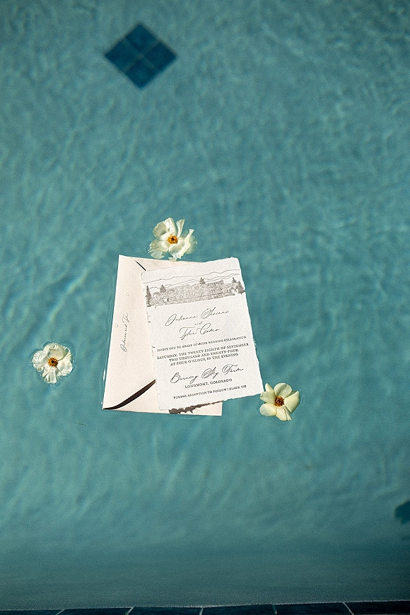 Wedding invitation suite with deckle edge wedding invitations, calligraphy card and venue sketch beside envelope on pool tile by rippling water