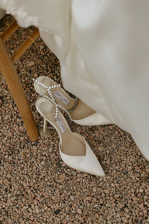 Bridal shoes, ivory wedding heels with pointed toes and rhinestone ankle straps, resting on gravel beside a wooden chair leg