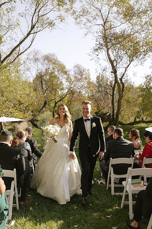 Wedding recessional as bride and groom walk the aisle, bride holding a white rose bouquet, sunlight on a tree-lined lawn with guests seated