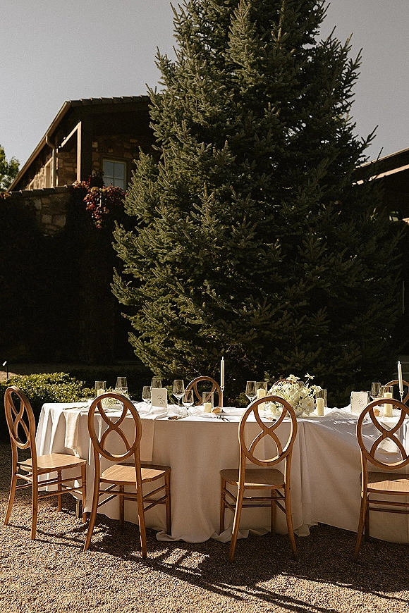 Reception tablescape with an outdoor reception table set in white linen, taper candles, wine glasses, and white florals by a stone building