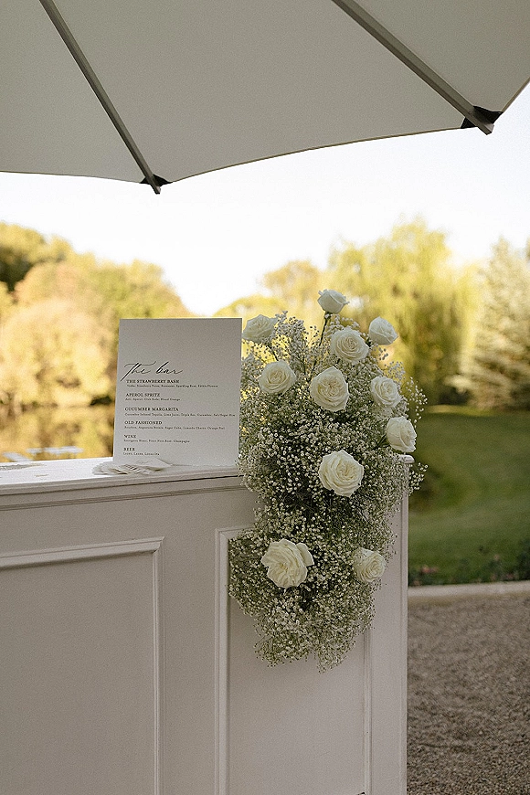 Wedding bar decor with a wedding bar menu sign and white rose accents on a white bar front, set on a garden lawn by trees