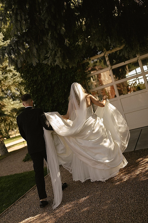 Wedding couple portrait of bride and groom walking away, bride lifting her dress train and veil as he helps on a sunlit garden path
