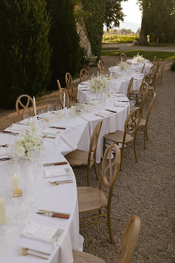 Reception tablescape with outdoor wedding dinner tables set on white linens, white floral centerpieces and taper candles in a hedge-lined gravel courtyard