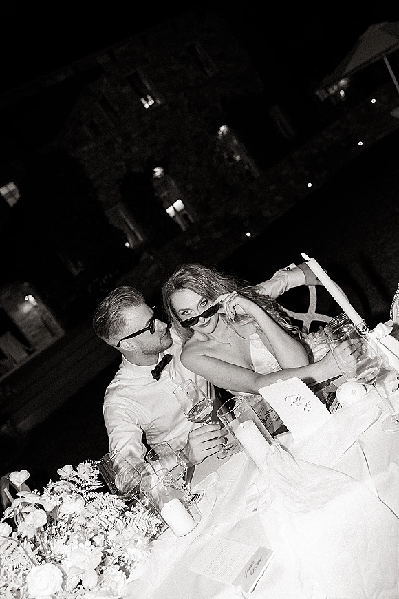 Reception couple portrait of bride and groom at table toasting in sunglasses amid candlelit florals and string lights in a garden night setting