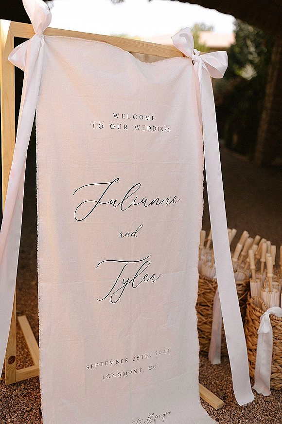Wedding welcome sign on a wooden easel, linen fabric with calligraphy and white ribbon bows, beside baskets and paper cones at sunset walkway