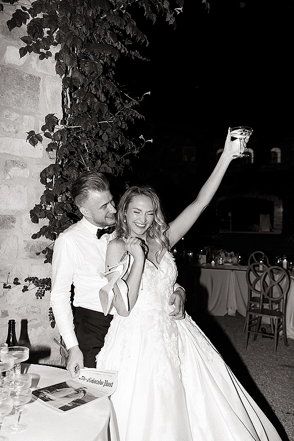 Reception couple moment in a black and white wedding photo, groom hugging bride as she raises a champagne coupe by ivy stone wall at night
