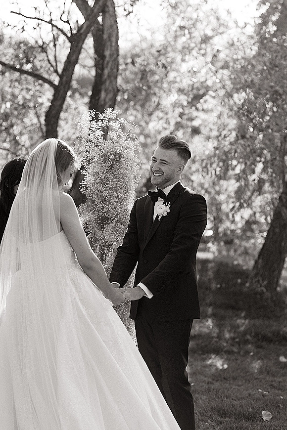 Ceremony moment of couple holding hands as groom smiles at bride, her veil trailing behind, in a sunlit garden setting