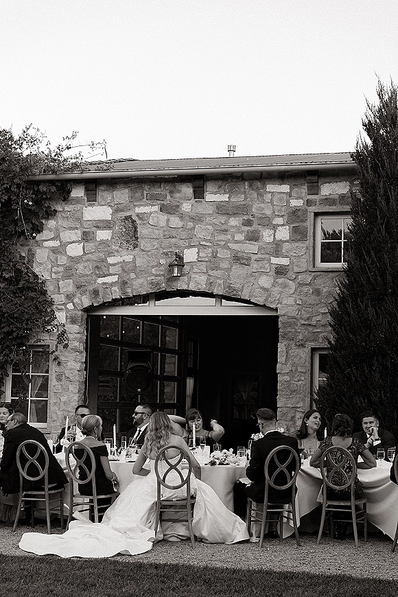Wedding reception table set with white linens, floral centerpiece and taper candles, beside a bride’s gown train on a stone villa lawn
