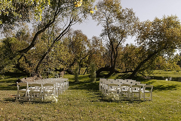 Ceremony setup with white folding chairs and aisle florals arranged for an outdoor wedding ceremony on a grassy lawn by a lake under trees