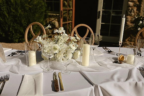 Reception tablescape with a white wedding tablescape, white floral centerpiece, candles, and place cards on an outdoor patio by glass doors